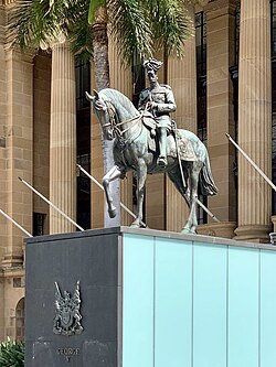 Equestrian statue in dark grey metal of George V in military dress uniform on a plinth of red granite outside a Classical building of red sandstone