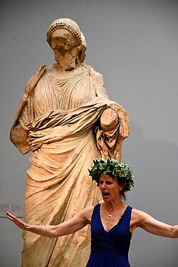 An actress performing a play. She wears an ivy wreath and stands in front of a statue of a woman from the Mausoleum at Halicarnassus (room 21, The British Museum, London)