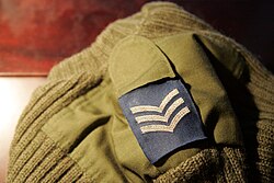 Shoulder mark of a contemporary British OG (Olive Green) pullover with RAF Sergeant insignia. The shoulder strap is attached with hook-and-loop fastener.