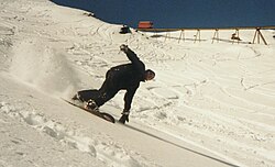 A snowboarder freeriding in deep snow. In the background, the Olympiabahn funicular built for the 1976 Olympics can be seen.