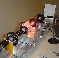 A helium–neon laser demonstration. The glow running through the center of the tube is an electric discharge. This glowing plasma is the gain medium for the laser. The laser produces a tiny, intense spot on the screen to the right. The center of the spot appears white because the image is overexposed there.