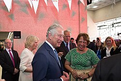 Charles in a blue suit smiles while interacting with a woman in a green dress at a public event. People surround them, capturing the moment with phones.