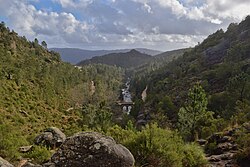 A wide view of a forested mountain valley in the Peneda-Gerês National Park under grey clouds, with rocky slopes on both sides, a stream running through the centre, and a small stone bridge partly hidden among the trees