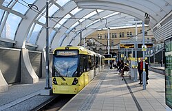 Two yellow trams sit beside one another on parallel tracks, with the one on the left slightly closer to the camera which is on the ground in the road. Both have their pantographs up which are touching contact wires. On either side there are people walking on the pavement.
