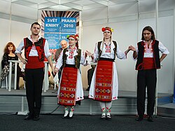 Bulgarian folk dancers in a national costume with embroidery on the penultimate row of the aprons showing the most spread Slavic cryptogram Bur[187] with a cross inside the rhombus representing the sun and spirals indicating rain,[188] which is similarly represented as the Rising Sun[189] decorative pattern of the Flag of Belarus. Similar carpet patterns appear on the Flag of Turkmenistan ultimately derived from ancient Persia.