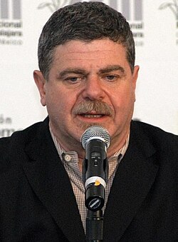 A 56-year-old man with dark gray hair talking into a microphone, looking at something beneath the camera