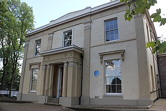 A small stucco building with sash windows. It is a cube shape with a slightly slanted roof and two storeys, as photographed from the ground in front. Trees are seen on either side of the building.