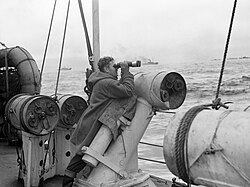 A lookout of a convoy escort, posing his binoculars on a depth charge thrower with which depth charges were launched to the sides of the escort