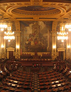 A view of a large, ornately-decorated room with several rows of curved desks, arranged in a semicircle. A large mural is visible on the wall at the far end of the room.
