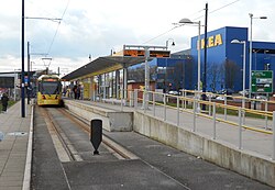 A tram terminus consisting of an island platform with a tram on the left hand side and a buffer just in front of the camera. The IKEA is visible in the background. The station is mostly built from clean concrete