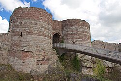 Two round towers of light yellow stone at the bottom and dark orangy stone at the top on either side of an arched entrance. A bridge leads from the entrance to allow access.