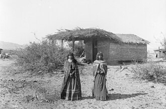 Two Mojave girls standing in front of a small dwelling with a thatched roof, 1900