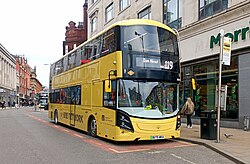 Three yellow buses line up such that the front one is just right-of centre and they are at an angle with the third one going offscreen. They are at a bus stop with tall buildings all around and someone waiting outside the first bus; the photo is taken from across the road.