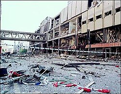 A street covered in debris. On one side is a building whose side is half-destroyed, leaving only the steel frame. The road is wet and there is crime scene tap across the centre of the photo.
