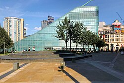 A triangular prism-shaped glass building as photographed from a short distance away, with trees lined in front