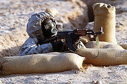 A Syrian soldier aims a Type 56 assault rifle from a foxhole during a military activity demonstration in Saudi Arabia in 1990. The soldier is wearing a Soviet-made Model ShMS nuclear-biological-chemical warfare mask.