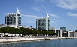 Photography of Parque das Nações, in Lisbon. Modern waterfront buildings, including two tall glass-and-concrete towers with sail-shaped white roofs, can be seen across the Tagus River under a blue sky with few thin white clouds. Trees, a riverside promenade, pedestrians, and nearby commercial buildings line the waterfront.