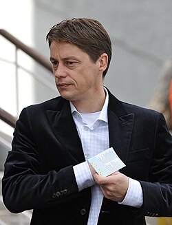 The upper body of a middle-aged white man with brown hair. He is wearing a light-coloured shirt with light blue pinstripes over a white T-shirt with a dark jacket on top. His right hand is inside his jacket and his left hand is holding a piece of paper.