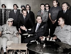 Three important seated men conferring. The first man from the left is wearing a checkered headdress, sunglasses and jodhpurs, the second man is wearing a suit and tie, and the third is wearing military uniform. Standing behind them are suited men.