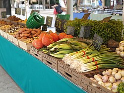 A stall at an outdoor market displaying pumpkins, sweet potatoes, onions, corn, herbs, and other vegetables arranged in wooden crates.