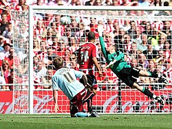 A football player in a red and white shirt and a football player in a light blue shirt are watching the ball approaching the goal, while the goalkeeper is diving to his right-hand side