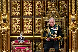 Charles seated on a throne during the 2022 state opening of the British Parliament. Next to him is the Imperial State Crown.
