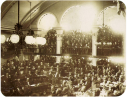 A photograph showing a large number of men seated on semi-circular tiers in a vaulted chamber as a large crowd looks on from an arcaded balcony