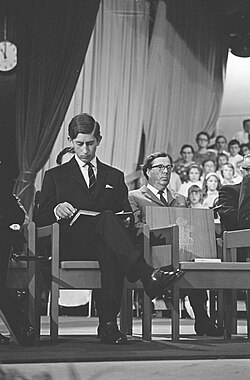 Charles in a suit sits on stage with a book, surrounded by others in similar attire. A choir is visible in the background.