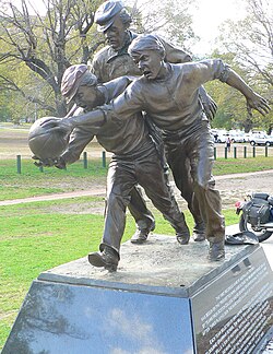 Statue next to the Melbourne Cricket Ground on the approximate site of the 1858 football match between Melbourne Grammar and Scotch College. Tom Wills is depicted umpiring behind two young players contesting the ball. The plaque reads that Wills "did more than any other person – as a footballer and umpire, co-writer of the rules and promoter of the game – to develop Australian football during its first decade."[9]