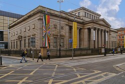 A neopalladian cuboid building as viewed centred on its corner from across the street. Halfway down the facade is a portico, and there is a central cuboid atrium that rises in the centre of the roof above the main roof height. There are regularly arranged sash windows in two storeys.