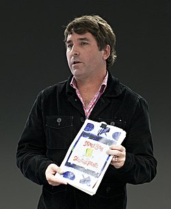 A man with short curly hair donning a black sweatshirt, showcases a bible of early concept art based on SpongeBob SquarePants as he speaks.