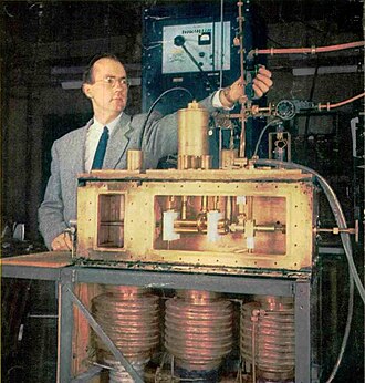 The first prototype ammonia maser in front of its inventor Charles H. Townes. The ammonia nozzle is at left in the box, the four brass rods at center are the quadrupole state selector, and the resonant cavity is at right. The 24 GHz microwaves exit through the vertical waveguide Townes is adjusting. At bottom are the vacuum pumps.