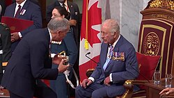 Mark Carney leans toward Charles, who is seated and wearing a suit adorned with military medals. A Canadian flag is visible in the background.