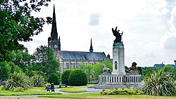 A church in the background-centre with its steeple on the left-hand side. On the right hand foreground a grand war memorial with a bronze statue on top of a large pillar. The foreground is set in a park with trees and paths