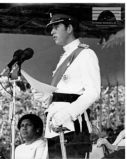 Charles in formal military attire stands at a podium outside, holding a document as he addresses an audience out of frame.