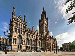 A grand Gothic Revival building as viewed from afar, with a Gothic Revival clock tower stretching high into the sky. Trees partially obstruct the view, and the sky is clear with some clouds.