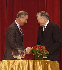 Charles warmly shake hands on stage with Scully, with a red curtain backdrop. A gold table in front holds a glass award and a floral arrangement.