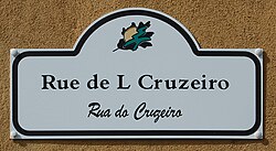 Photograph of a white street sign mounted on a tan wall reads in Mirandese "Rue de L Cruzeiro" with "Rua do Cruzeiro" written in Portuguese below in script, and a small leaf logo at the top.