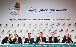 Several people dressed in formal attire sit behind a bench. Behind them, there are repeated graphics representing Rio 2016.