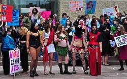 Five people dressed in different historic costumes, many with legs, arms and stomachs bared, hold hands in the foreground. Behind them, protestors hold handmade signs like "A Dress is not a Yes", "There's no shaming this slut", and a drawing of Ariel with the caption "She's not asking for it!"