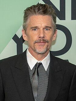 A Caucasian male with gray hair, wearing a two-piece dark gray suit with a gray and white shirt