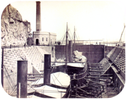 A photograph with two steamships resting in a dewatered drydock with a building housing the engine for operating the lock's gates in the background