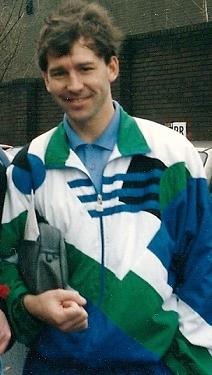 A smiling man with dark hair wearing a white, green, and blue tracksuit top over a blue shirt. He is holding a washbag under his right arm.