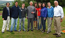 Charles, wearing a suit, stands with a group of eight people casually dressed, smiling on a grassy field. Trees are visible in the background.