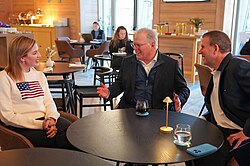 A group of professionally dressed people seated around a round table in a modern, warmly lit lounge, engaged in conversation and smiling. David Zaslav sits at the center, gesturing while speaking, with a woman in a white sweater featuring an American flag seated to his left and a man in a dark blazer leaning in from his right. Drinks rest on the table, while other guests and soft ambient lighting fill the background.