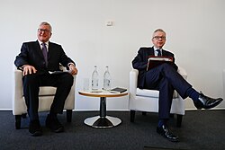 Two men seated in armchairs beside a small table during a discussion