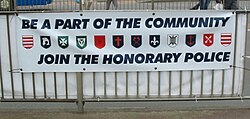 A recruiting banner for the Honorary Police showing the arms of each parish: (from left to right) Grouville, St Brelade, St John, Trinity, St Saviour, St Ouen, St Helier, St Mary, St Lawrence, St Clement, St Peter, St Martin