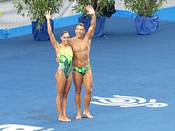 A mixed-sex pair, participating in World Aquatics Championships of synchronised swimming, waves to the crowd before diving into water.