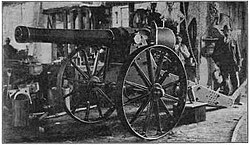 The "Long Cecil" gun in the workshops of the De Beers company showing a large gun on a carriage with a workman casually looking on in the background.