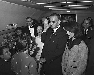 Johnson is sworn in as president aboard Air Force One at Dallas Love Field two hours and eight minutes following Kennedy's assassination as Mrs. Johnson and Mrs. Kennedy look on.